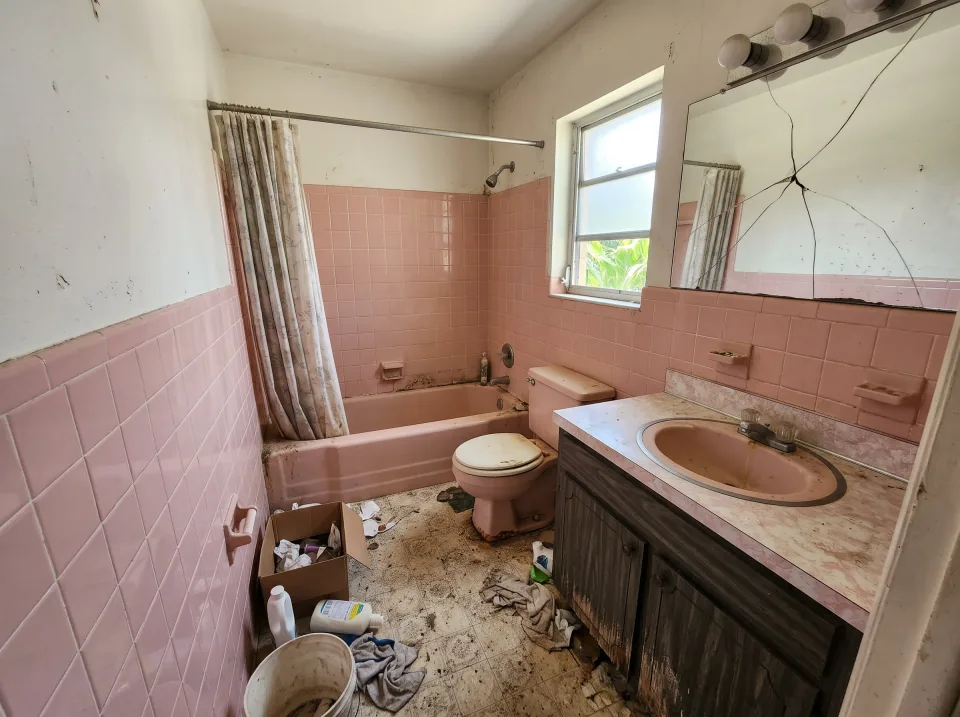Illustration: a dated bathroom in a 1970s Broward ranch house: pink tile walls, a pink tub, a vanity with a cracked mirror, a toilet, a small frosted window.