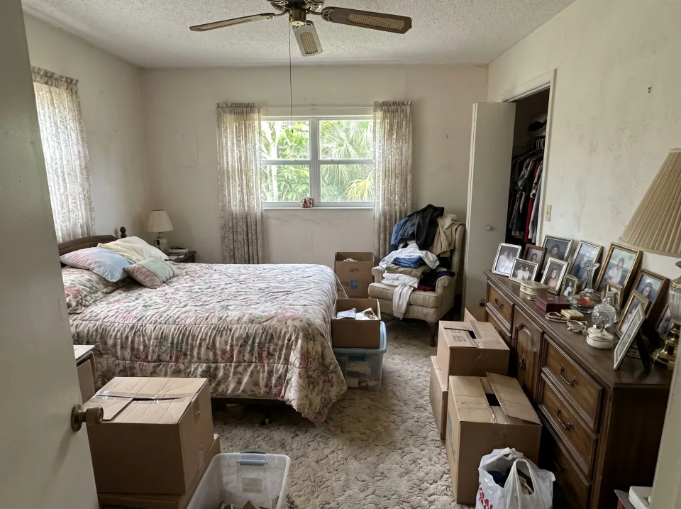 Illustration: a bedroom in an older South Florida home after a family member passed: a bed with a floral spread, a heavy dresser covered in framed photos and knickknacks, stacked boxes, clothes on a chair, lace curtains.