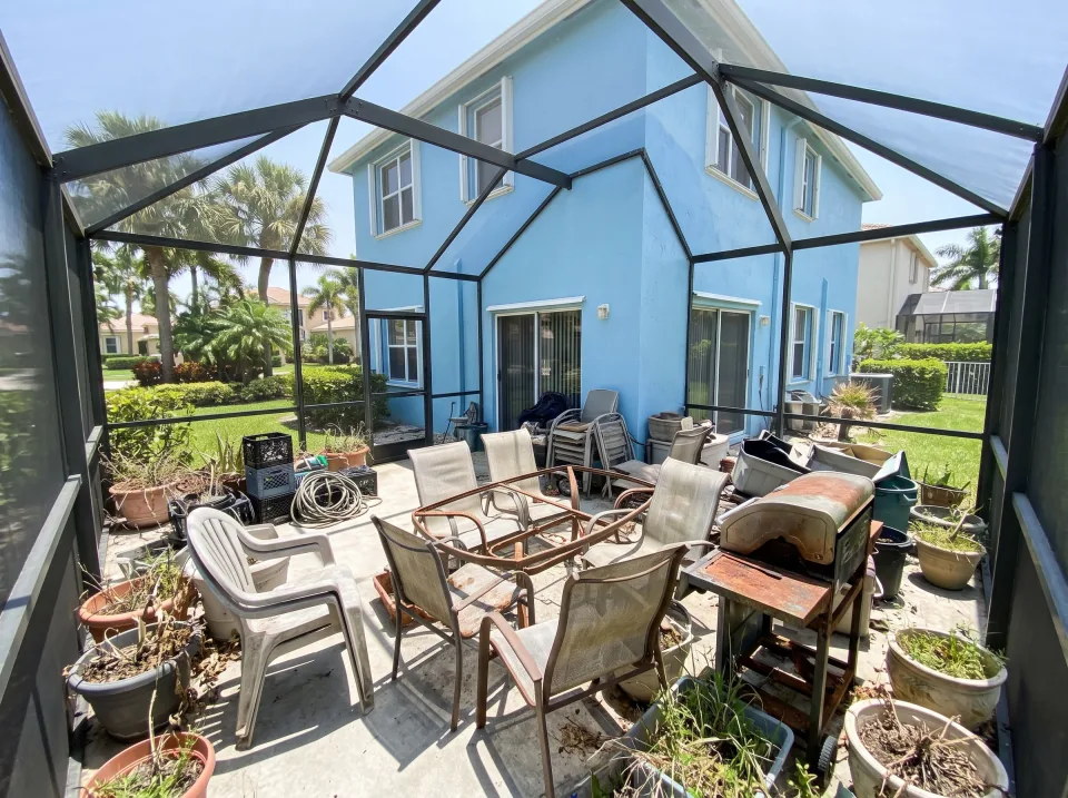 Illustration: a screened patio crowded with faded patio chairs, a broken table, an old grill and planters at a sky blue two-story home in an Atlantis gated community with manicured landscaping, flat white roof, clear midday sun