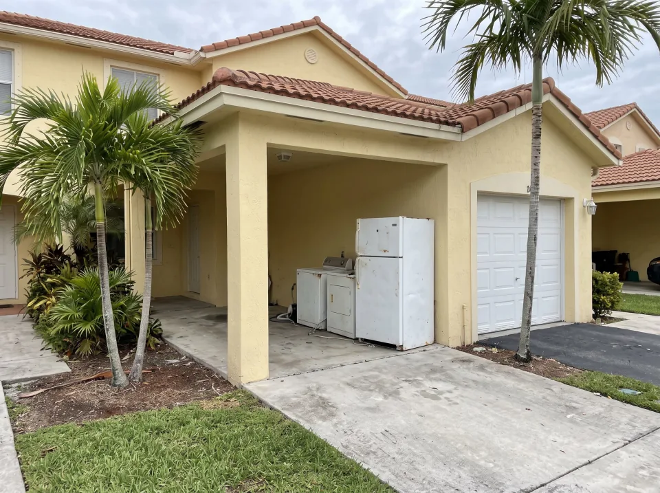 Illustration: a cluttered single-car garage in a Florida townhome with a broken chest freezer, old lawn mower, rusty tool shelves overflowing with junk, a stained mattress leaning on the wall and a pile of black trash bags.