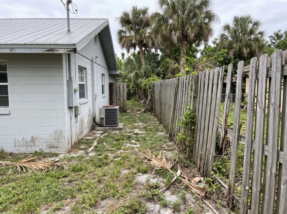 Illustration: a leaning, weathered wooden privacy fence with missing boards along the side yard at a light gray single-story Gulf Stream concrete block home with a standing-seam metal roof and palm trees, soft overcast light