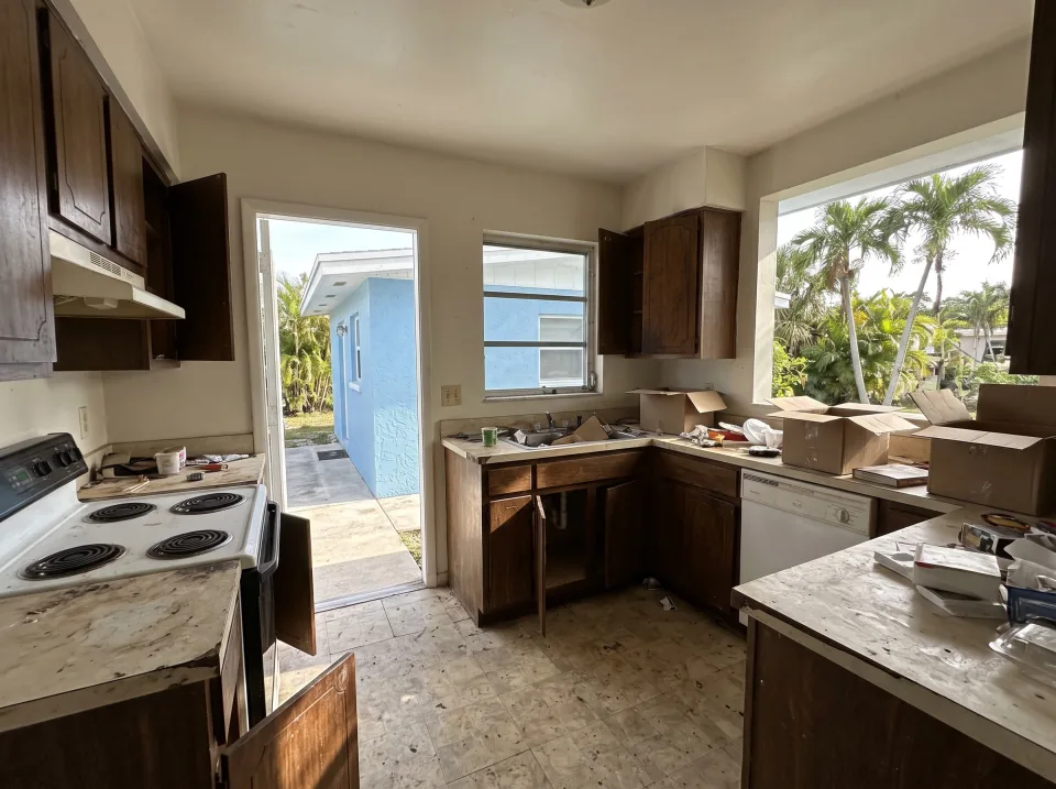 Illustration: a dated kitchen with old wood cabinets, laminate counters and a white stove at a sky blue single-story Pembroke Pines concrete block home with a flat white roof and palm trees, bright morning light