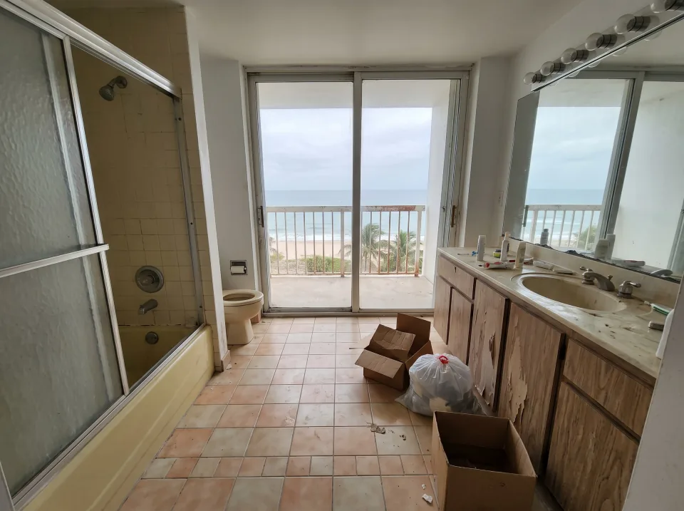 Illustration: a dated tiled bathroom with an old tub, toilet and vanity inside a Lauderdale-by-the-Sea beachside condo unit with ocean view through sliding glass doors, soft overcast light