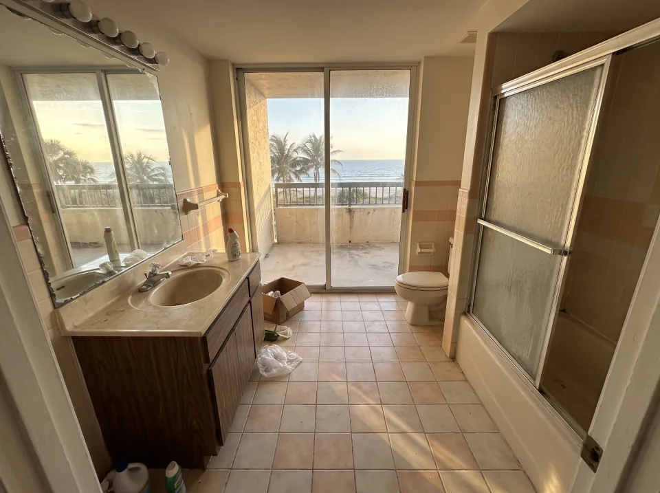 Illustration: a dated tiled bathroom with an old tub, toilet and vanity inside an Ocean Ridge beachside condo unit with ocean view through sliding glass doors, warm late-afternoon light