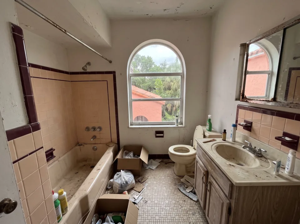 Illustration: a dated tiled bathroom with an old tub, toilet and vanity at a coral pink historic Opa-locka home with arched windows, terracotta tile roof and big shade trees, soft overcast light