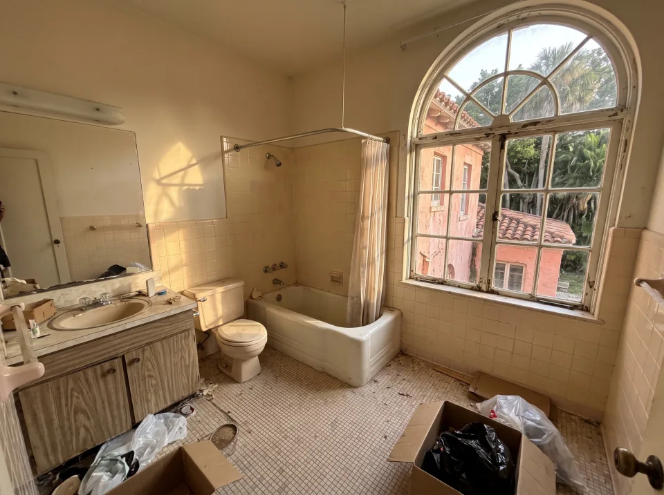 Illustration: a dated tiled bathroom with an old tub, toilet and vanity at a coral pink historic Palm Beach home with arched windows, barrel tile roof and big shade trees, warm late-afternoon light