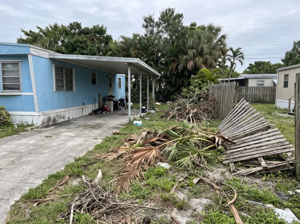 Illustration: a side yard full of storm-fallen branches, palm fronds and an old fence section at a sky blue mobile home in a Pembroke Park park with a carport and small yard, soft overcast light
