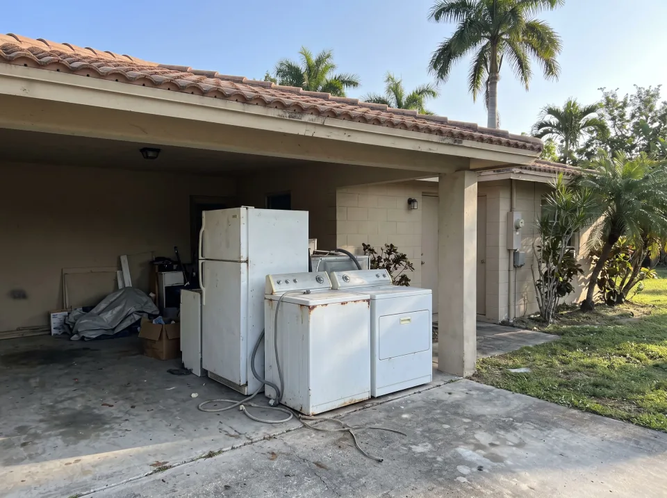 Illustration: an old refrigerator, a washer and a dryer pushed together in the carport at a sand beige single-story Royal Palm Beach concrete block home with a barrel tile roof and palm trees, bright morning light