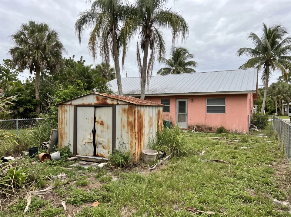 Illustration: a rusted metal garden shed with dented doors in the backyard at a coral pink single-story Weston concrete block home with a standing-seam metal roof and palm trees, soft overcast light