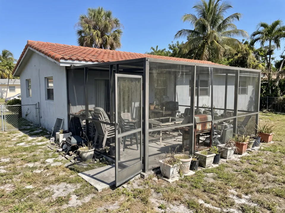 Illustration: a screened patio crowded with faded patio chairs, a broken table, an old grill and planters at a light gray single-story West Miami concrete block home with a barrel tile roof and palm trees, clear midday sun
