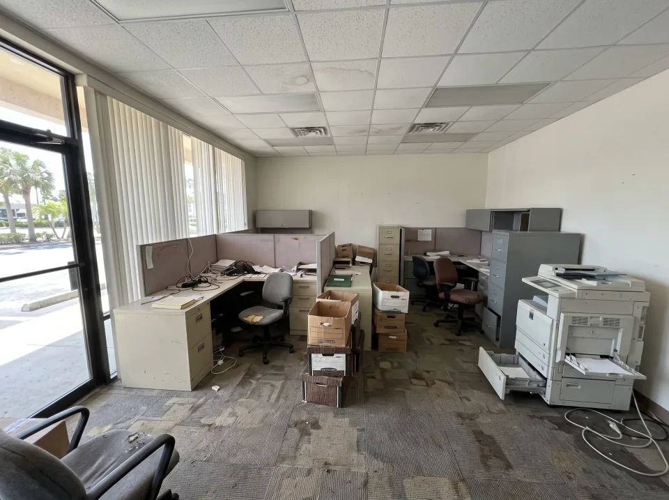 Illustration: a small commercial office suite in a Florida strip-mall building with old cubicle desks, office chairs, filing cabinets, boxes of paper and a dead copier, carpet tile floor.